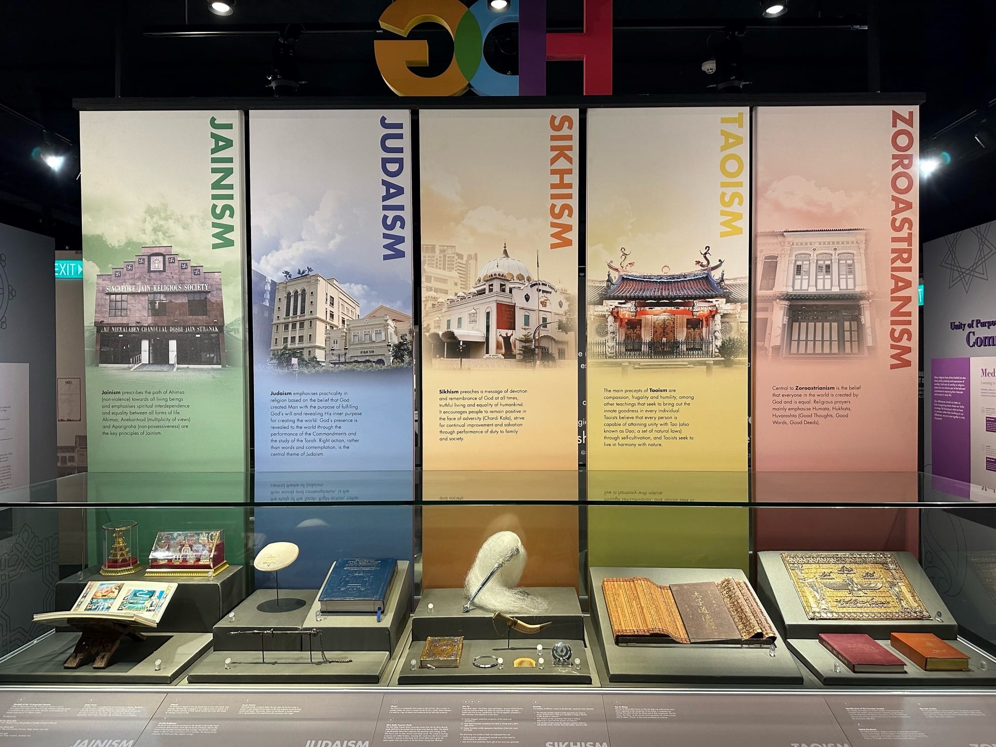 Museum exhibit with colorful panels on Jainism, Judaism, Sikhism, Taoism, and Zoroastrianism, with artifacts in a display case below.